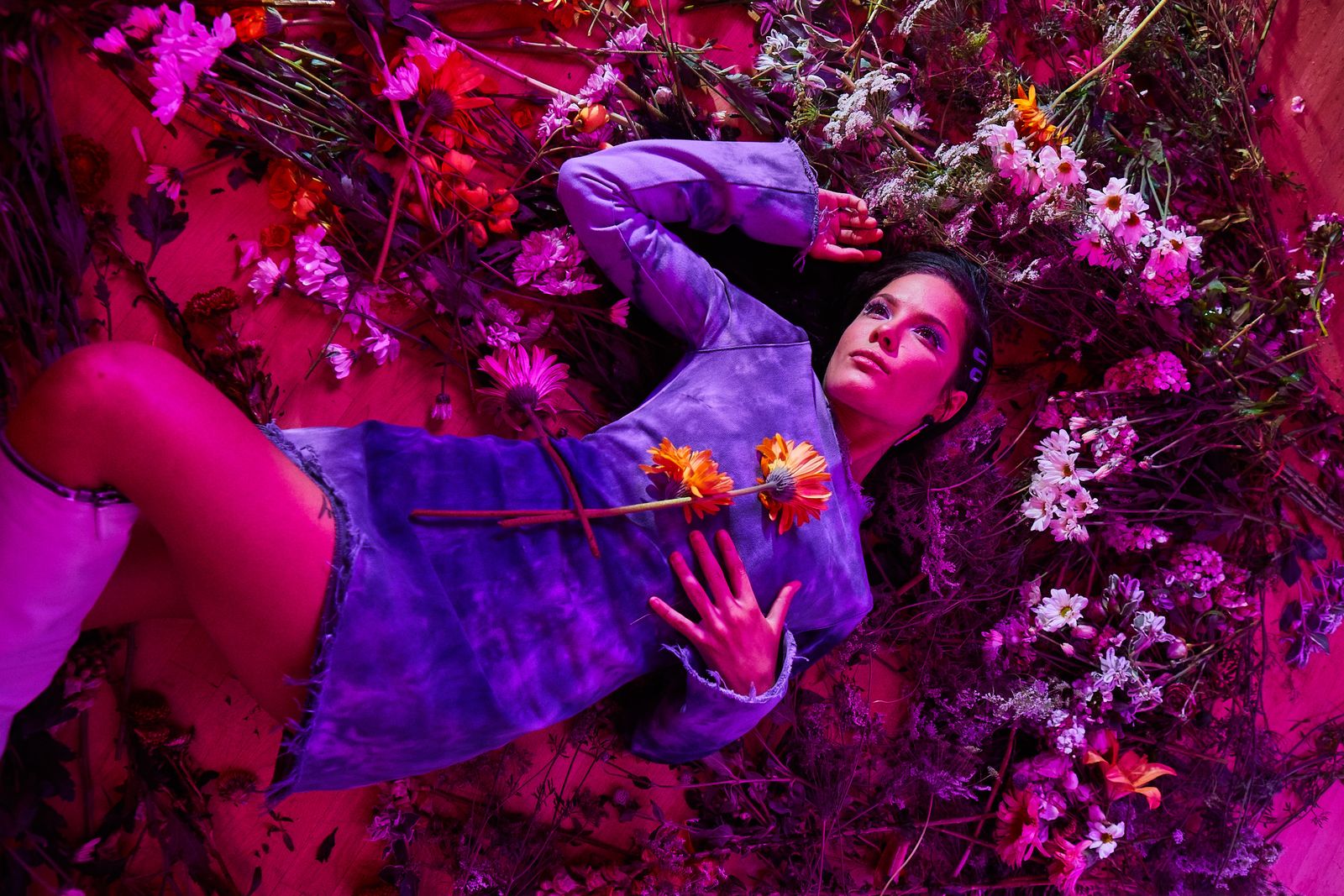 Woman in a purple tie-dye dress lying on a bed of flowers under magenta light